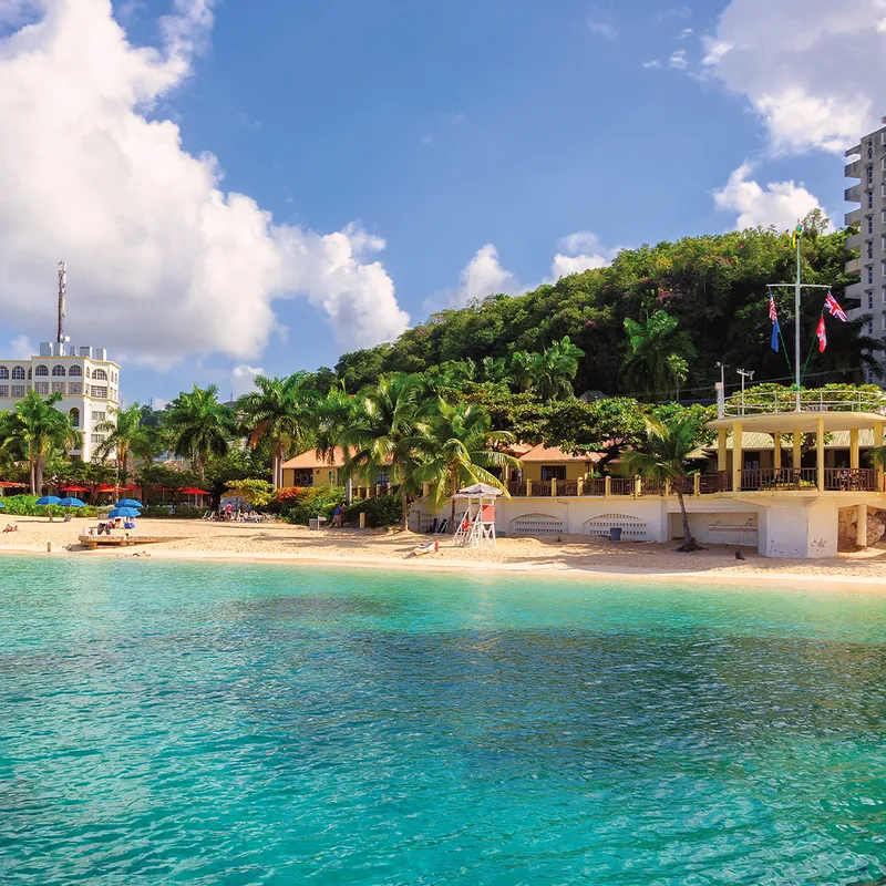 Beachfront scene with turquoise water, sandy shore, palm trees, and colorful buildings in Jamaica