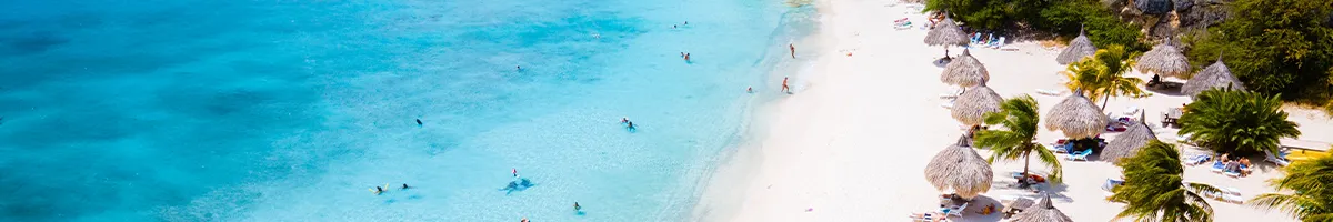 Aerial view of a tropical beach with turquoise water, people swimming, and sunbathers under thatched umbrellas surrounded by palm trees.